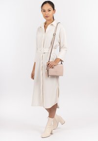Abito lungo color crema con una cintura, abbinato a una piccola borsa beige con un pon-pon. La modella indossa stivaletti alla caviglia eleganti e di colore chiaro.
