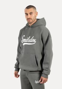 Szara bluza z kapturem z miękkiego materiału, z przednią kieszenią, z białym napisem "Smilodox" wyraźnie wyeksponowanym na piersi. Model pokazany w ubraniu.