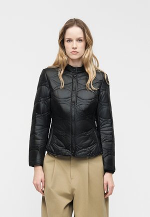 Jeune femme aux longs cheveux blonds portant une veste matelassée noire et un pantalon large beige, debout devant un fond clair uni.