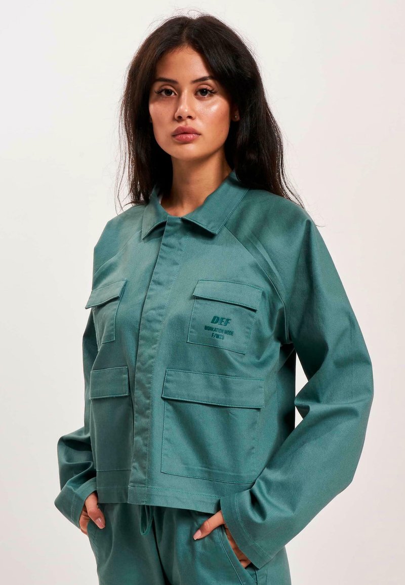 Türkisfarbene, kurz geschnittene Jacke aus glattem Stoff, mit Kragen, zwei vorderen Taschen und dezenter Branding auf der Brust. Lässige Passform.