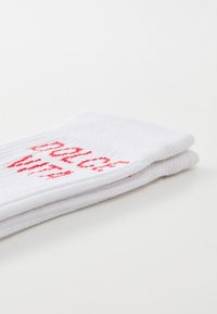 Biele bavlnené ponožky s červeným vyšitým textom. Dizajn zahŕňa rebrovanú textúru a prehnutý lem na vrchu.