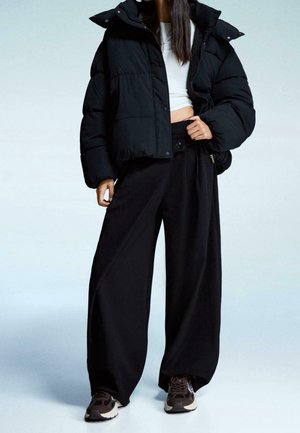 Vrouw draagt een zwarte donsjas, brede zwarte broek, wit cropped top en zwarte sneakers, staand tegen een eenvoudige lichte achtergrond.