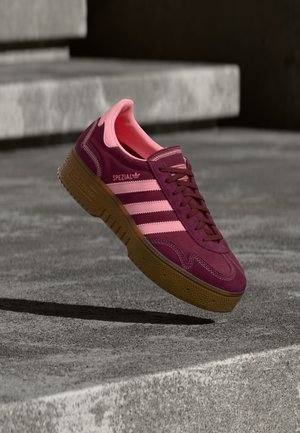 Bordeauxrode suède sneaker met roze accenten, voorzien van drie strepen, een getextureerde gumzool en een versterkte neuskap. Strak ontwerp, laag profiel.