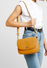 Kabelka na rameno z tan kůže s klopou, zdobená zlatým logem a hardwarem. Nastavitelný popruh a hladký povrch.