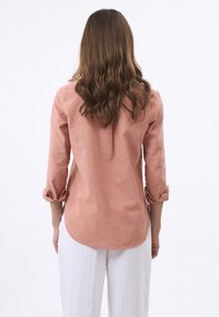 Camicia di lino rosa con maniche a tre quarti, polsini arrotolati e orlo curvo. Il tessuto ha una trama morbida e una sottile lucentezza.