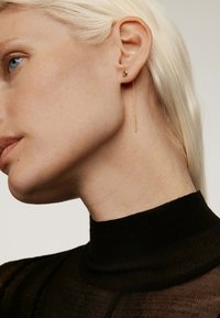 Pendientes de oro con un pequeño stud en forma de bola y una larga cadena delgada. La modelo tiene el cabello rubio corto y liso y lleva una blusa negra ajustada.