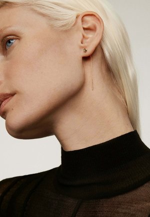 Pendientes de oro con un pequeño stud en forma de bola y una larga cadena delgada. La modelo tiene el cabello rubio corto y liso y lleva una blusa negra ajustada.