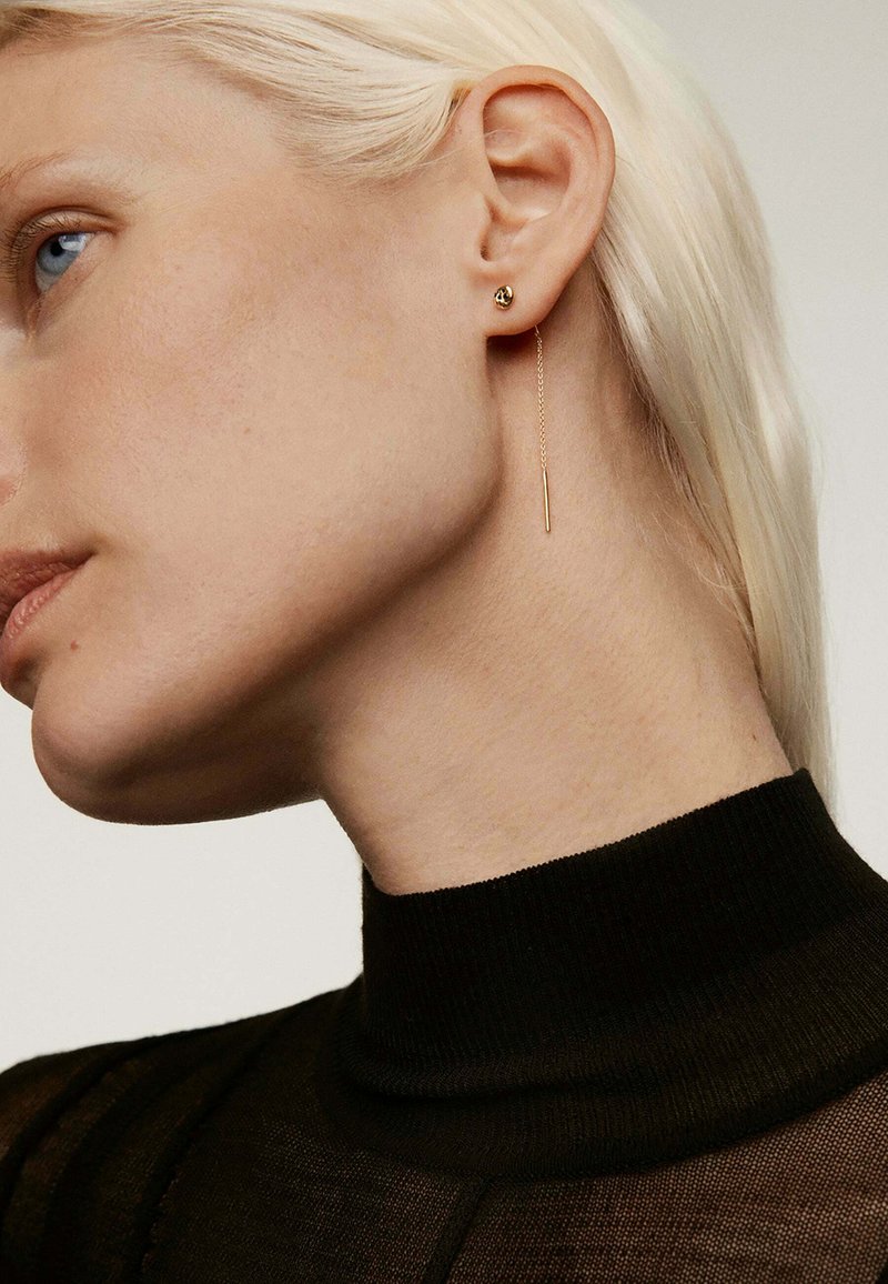 Pendientes de oro con un pequeño stud en forma de bola y una larga cadena delgada. La modelo tiene el cabello rubio corto y liso y lleva una blusa negra ajustada.