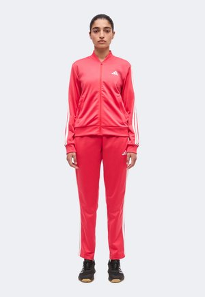 Naine punases Adidas dressipluus valgete triipude ja mustade tossudega, seisab tavalise heleda tausta ees.
