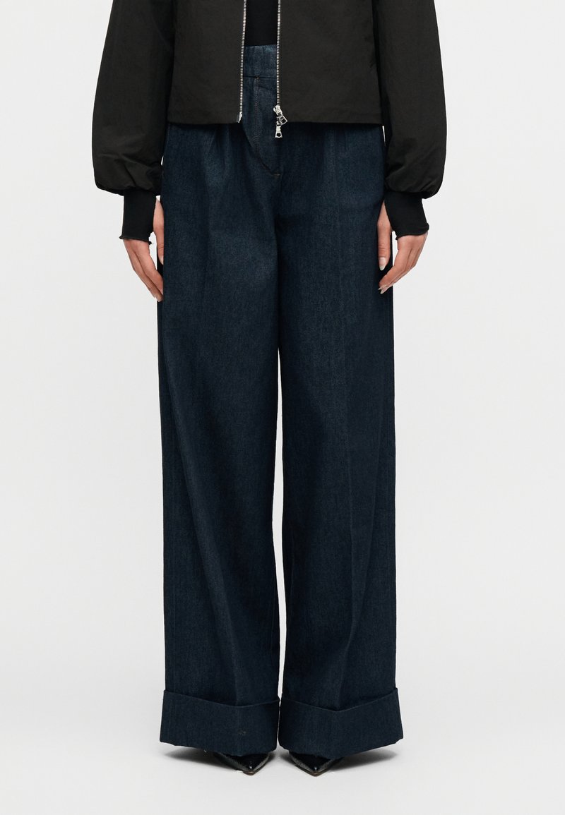 Γυναίκα που φοράει σκούρο παντελόνι με φαρδιά μπατζάκια και μανσέτες, καθώς και μαύρο μακρυμάνικο σακάκι με εμφανές φερμουάρ, στέκεται μπροστά σε ένα απλό φόντο.