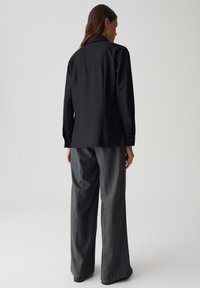 Maglietta nera a maniche lunghe con una vestibilità ampia e colletto classico, abbinata a pantaloni larghi grigi a righe con dettagli dei bottoni visibili sulle maniche.