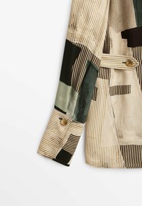 Veste multicolore arborant des motifs abstraits, des rayures et des patchs dans des tons de vert, noir et beige ; un bouton décoratif près du poignet.