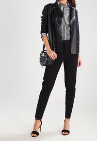 Donna con giacca di pelle nera sopra una maglietta a strisce bianche e nere, pantaloni neri, tacchi alti neri, che tiene in mano una borsa rotonda nera.