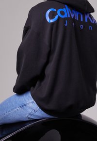 Sudadera con capucha negra con un logo azul brillante de "Calvin Klein Jeans" en la espalda, combinada con vaqueros de mezclilla azul claro, sentada sobre una superficie negra y brillante.