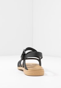 Svart sandal med flera justerbara remmar, en tan gummisula, texturerad fotbädd och spänne på sidan. Lämplig för avslappnad användning.