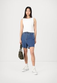 Un haut sans manches blanc, un short en denim bleu taille haute, des baskets montantes blanches et un sac en tissu vert. Textures et détails de design minimalistes.