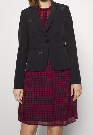 Blazer noir avec fermeture à bouton sur le devant et ornements en forme d'étoile ; superposé sur une robe à motifs pied-de-poule rouge et noir, comportant un décolleté à volants.