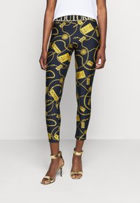 Svarta leggings med ett mönster av guldkedjor och accessoarer, med en texturerad midja. Kombineras med guldiga sandaletter med hög klack och remmar.