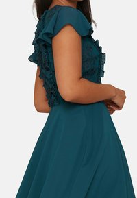 Tealfärgad klänning med en figursydd livstycke prydd med intrikata blommor i spets och volangärmar, flödande kjol och slät tyg med en lätt lyster.