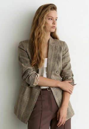 Junge Frau mit langen blonden Haaren, die einen beigen karierten Blazer, ein weißes Oberteil und braune Hosen trägt, steht mit verschränkten Armen und schaut seitlich.