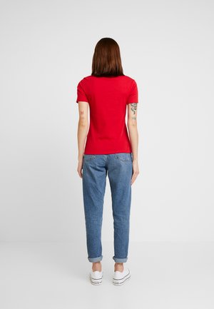 Kvinde med skulderlangt brunt hår iført en rød kortærmet skjorte, blå opkrogede jeans, hvide sneakers, stående med ryggen mod fronten.