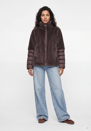 Frau trägt eine braune Fleecejacke mit gesteppte Ärmeln, helle Jeans mit weitem Bein und braune Sneakers und steht vor einem einfarbigen weißen Hintergrund.