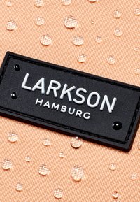 Tessuto color pesca con gocce in rilievo, caratterizzato da un'etichetta rettangolare nera in rilievo con la scritta "LARKSON HAMBURG" in lettere bianche.