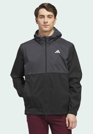 Junger Mann trägt eine schwarz-dunkelgraue Kapuzenjacke mit Reißverschluss und weißem Logo auf der Brust, steht mit Händen in den Taschen.