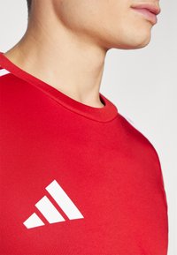 Red athletic t-shirt featuring a round neckline, short sleeves, and a prominent white Adidas logo on the chest. Smooth fabric texture.