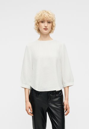 Personne aux cheveux blonds et bouclés portant un chemisier blanc texturé à manches ballon et un pantalon en cuir noir, debout devant un fond uni.