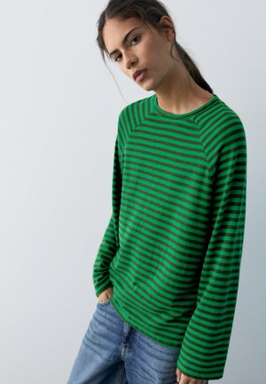 Jonge vrouw met donker haar in een paardenstaart, gekleed in een groen-bruin gestreept shirt met lange mouwen en een blauwe spijkerbroek, staand met handen in de zakken.