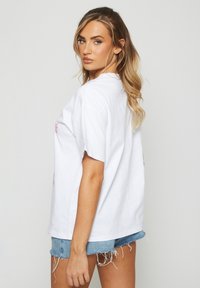 Kvinna med långt blont hår klädd i en vit oversized T-shirt och slitna denimshorts, som tittar över sin axel mot en enkel bakgrund.