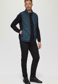 Gep quilteter, marineblauer Pufferweste mit hohem Kragen, schwarzem Reißverschluss, Seitentaschen, getragen über einem langärmeligen schwarzen Oberteil und schwarzen Hosen, kombiniert mit schwarzen Sneakers.