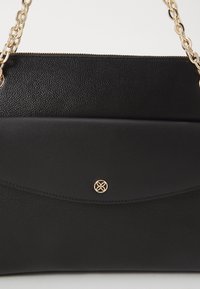 Borsa nera con pelle testurizzata e un pannello frontale liscio e curvo. Caratterizzata da una tracolla in catena dorata e un emblema decorativo in oro.