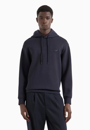 Sudadera con capucha de color azul marino en material suave, con un bolsillo tipo canguro en la parte frontal, capucha ajustable mediante cordón y emblema en el lado izquierdo del pecho.