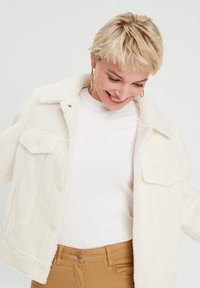 Veste en fausse fourrure blanche avec un col, des manches longues, deux poches poitrine et une coupe décontractée, portée sur un haut blanc.