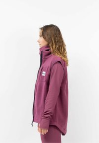 Fleece vest in diep paars, met een hoge kraag, ritssluiting aan de voorkant en zijzakken. Draagt over een lange mouw, aansluitend top in een bijpassende kleur.