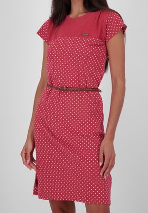 Kobieta w czerwonej sukience do kolan w białe grochy, z krótkimi rękawami i cienkim brązowym plecionym paskiem w talii, stojąca na jednolitym tle.