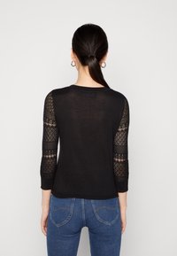 Schwarzer Pullover mit dreiviertellangen Spitzenärmeln, Rundhalsausschnitt und geripptem Saum. Getragen mit blauen Jeans, die die Gesäßtaschen zeigen.