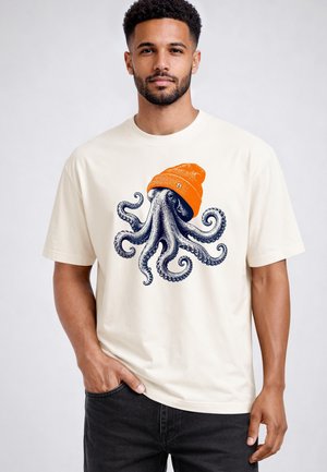 Mann mit einem hellfarbenen T-Shirt mit einem detaillierten Grafik eines Oktopus, der eine orange Mütze trägt, steht mit einer Hand in der Tasche.