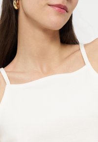 Γυναίκα που φοράει λευκό αμάνικο τοπ με λεπτούς ιμάντες, χρυσό κρίκο σκουλαρίκι και ροζ κραγιόν, με φόντο λευκό.