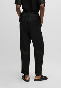 Czarne spodnie o luźnym kroju, z elastycznym pasem, bocznymi kieszeniami i subtelnym połyskiem; w połączeniu z czarnymi klapkami typu slip-on.