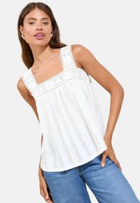 Haut sans manches blanc en tissu texturé, avec un décolleté carré, des détails brodés et un design plissé. Associé à un jean bleu.