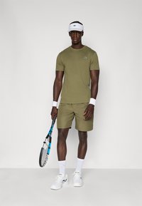 Ελαιώδη πράσινη αθλητική μπλούζα και σορτσάκι, λευκά αθλητικά παπούτσια, βραχιόλια και κορδέλα, κρατώντας μια τενιστική ρακέτα με μπλε λεπτομέρειες.