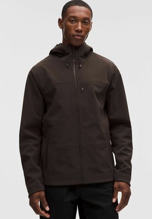 Hombre con chaqueta marrón oscura con cremallera y capucha, de pie frente a un fondo claro y simple.