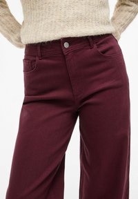 Jeans ampi in bordeaux con vita alta, tasche frontali e dettaglio in bottone argentato, realizzati in morbido denim testurizzato.