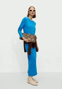 Niebieska sukienka z długim rękawem i beżowe botki za kostkę, trzymająca torebkę w leopardzi wzór, stylizowana z jasnymi okularami przeciwsłonecznymi i brązowym swetrem.