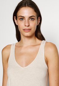 Beige tank top with a deep V-neckline, made from a soft, light-textured fabric. Features wide shoulder straps and a relaxed fit.