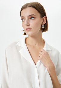 Blusa bianca a trama con colletto a V, chiusura a bottoni e vestibilità ampia. Il modello ha i capelli corti e indossa orecchini rotondi in argento.