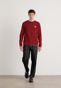Rode langemouwen shirt met ribbelstructuur, witte kraagdetail, gecombineerd met donkergrijze losse broek en zwarte schoenen.